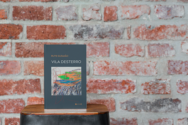 Imagem do livro "Vila desterro" de Rute Gusmão sobre mesa de madeira e fundo de tijolos vermelhos