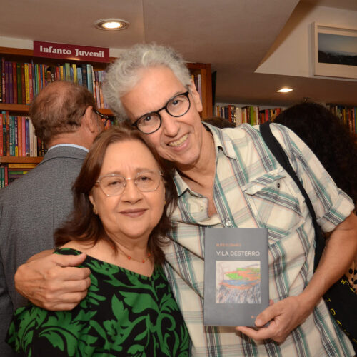 Rute posando em foto com Amador Perez segurando livro "Villa Desterro". Rute usa blusa preta com detalhes em verde e Amador usa blusa xadrez com fundo branco